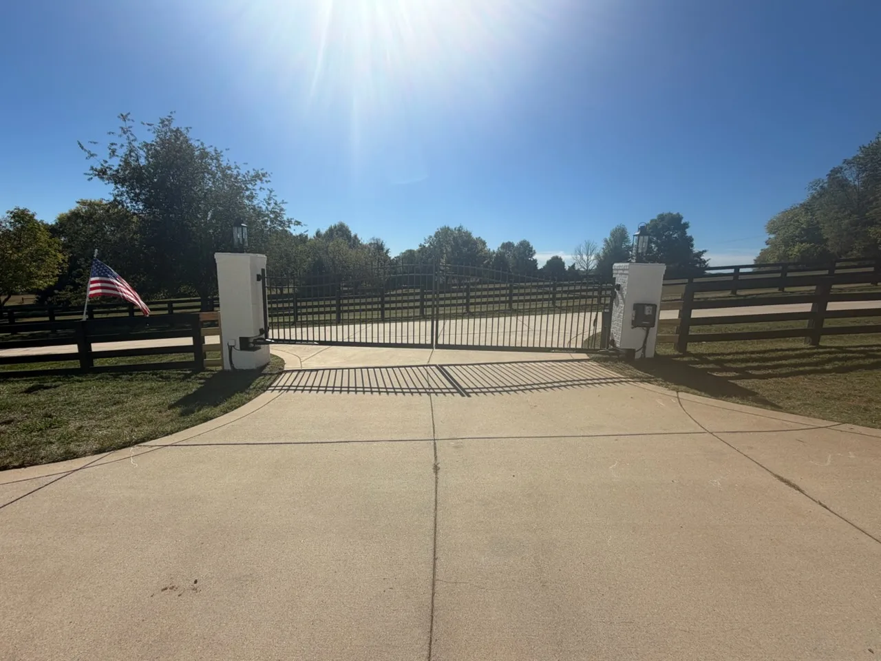 Black arched spear-top double swing gate closed across a concrete driveway between white painted brick columns with coach lanterns, gate operators on both columns and black four-board horse fence on either side