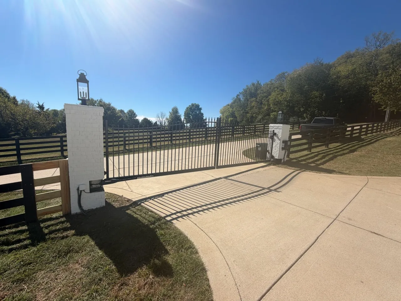Swing gate operator arm and weatherproof control box on a white brick column with a copper lantern, opening a black arched spear-top driveway gate beside a black board fence