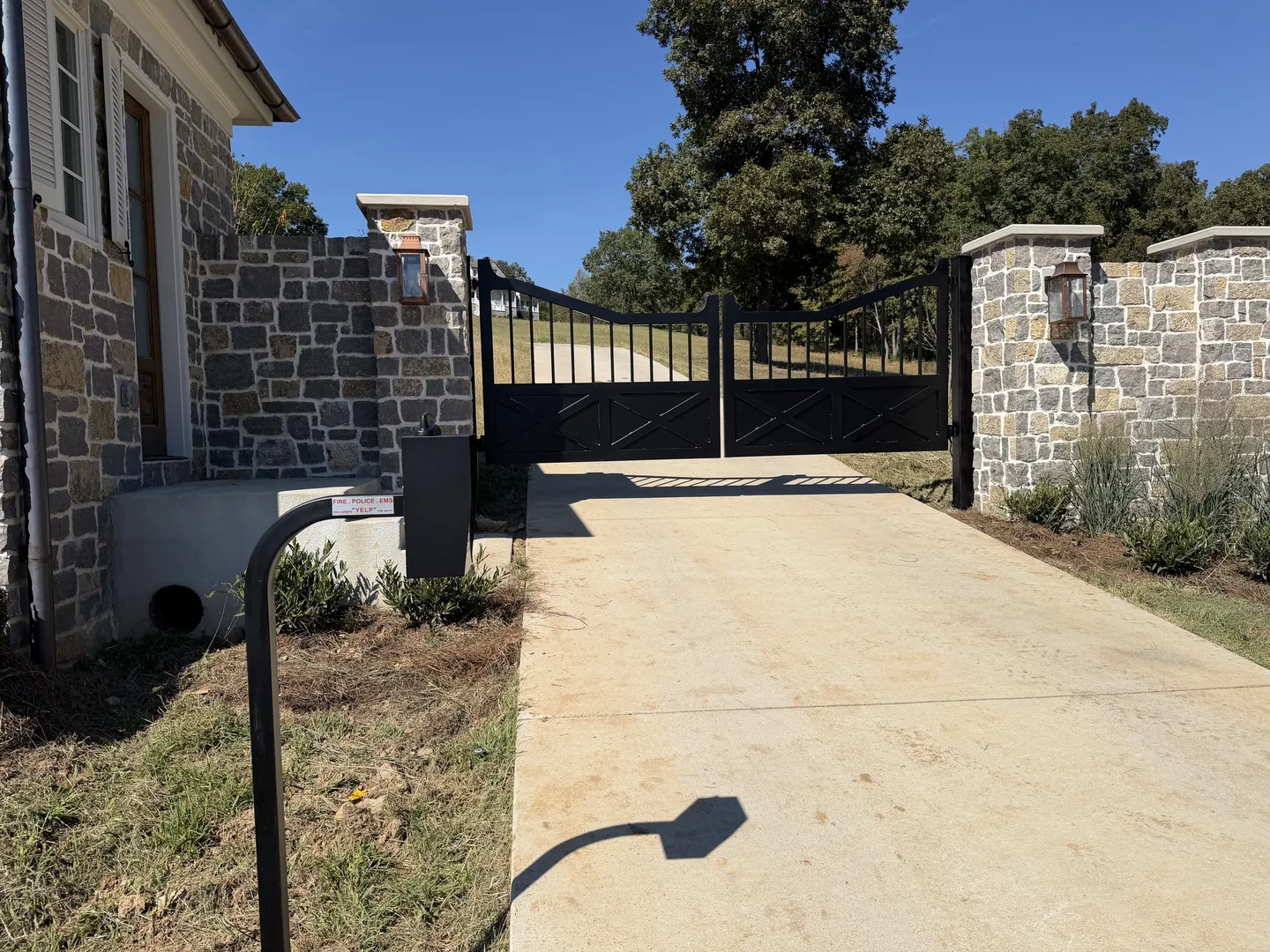 Black access control pedestal and emergency responder entry sign beside the stone column of an arched X-brace driveway gate, with a copper lantern mounted above