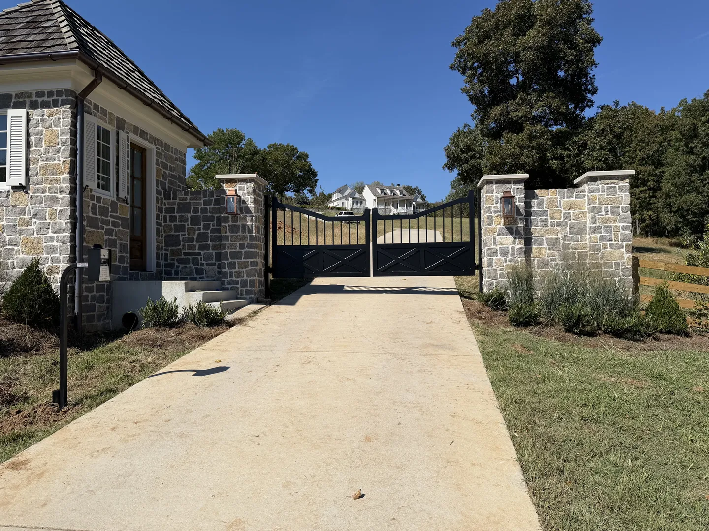 Black arched double swing gate with X-brace lower panels between stacked stone columns, a matching stone gatehouse at left and a white farmhouse on the hill beyond