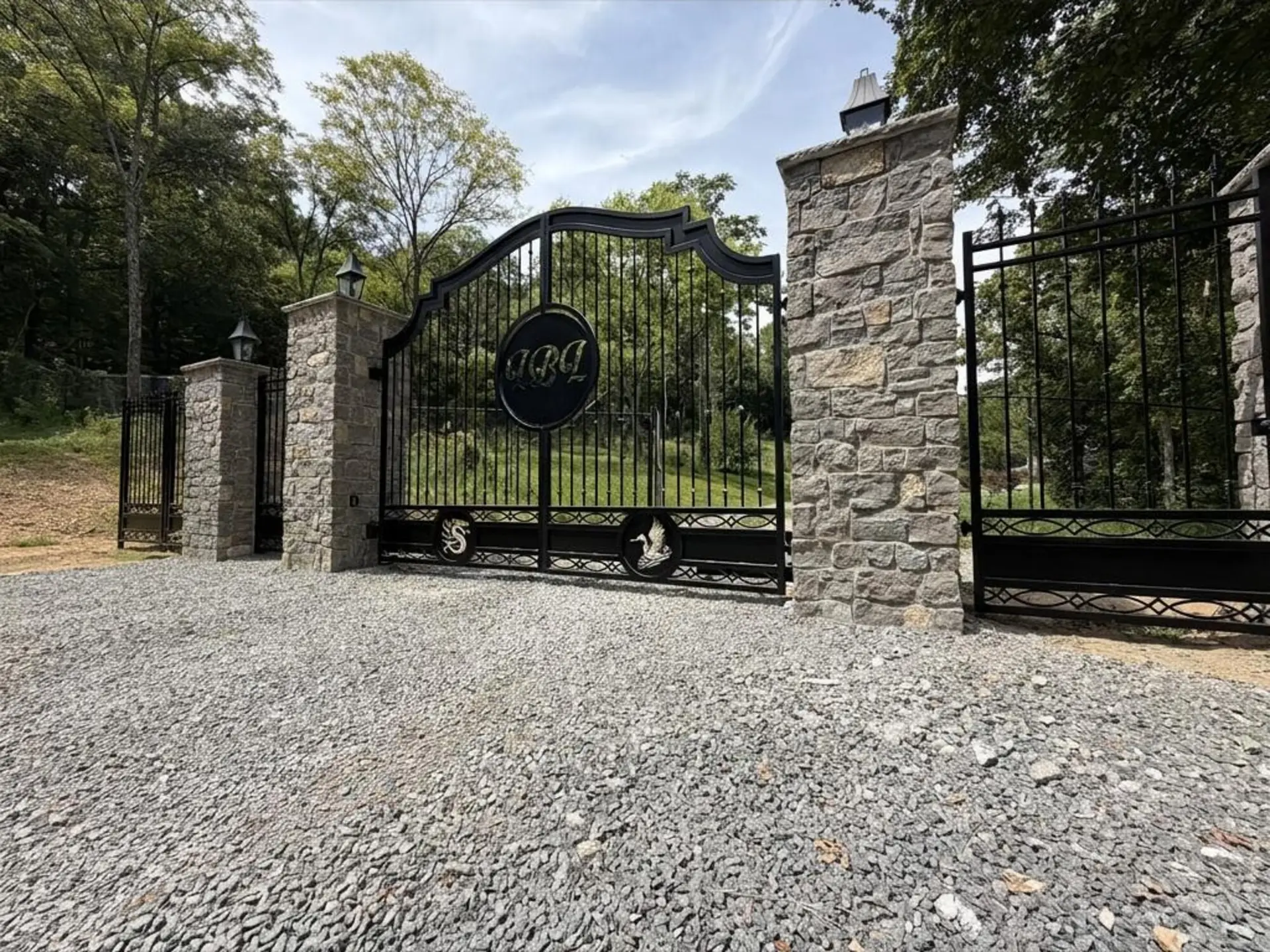 Custom ornamental iron driveway gate between stone columns on a clean gravel entrance